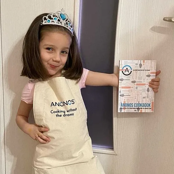 A young child wearing an Anonos 'Cooking without the drama' apron and holding the Anonos Cookbook.